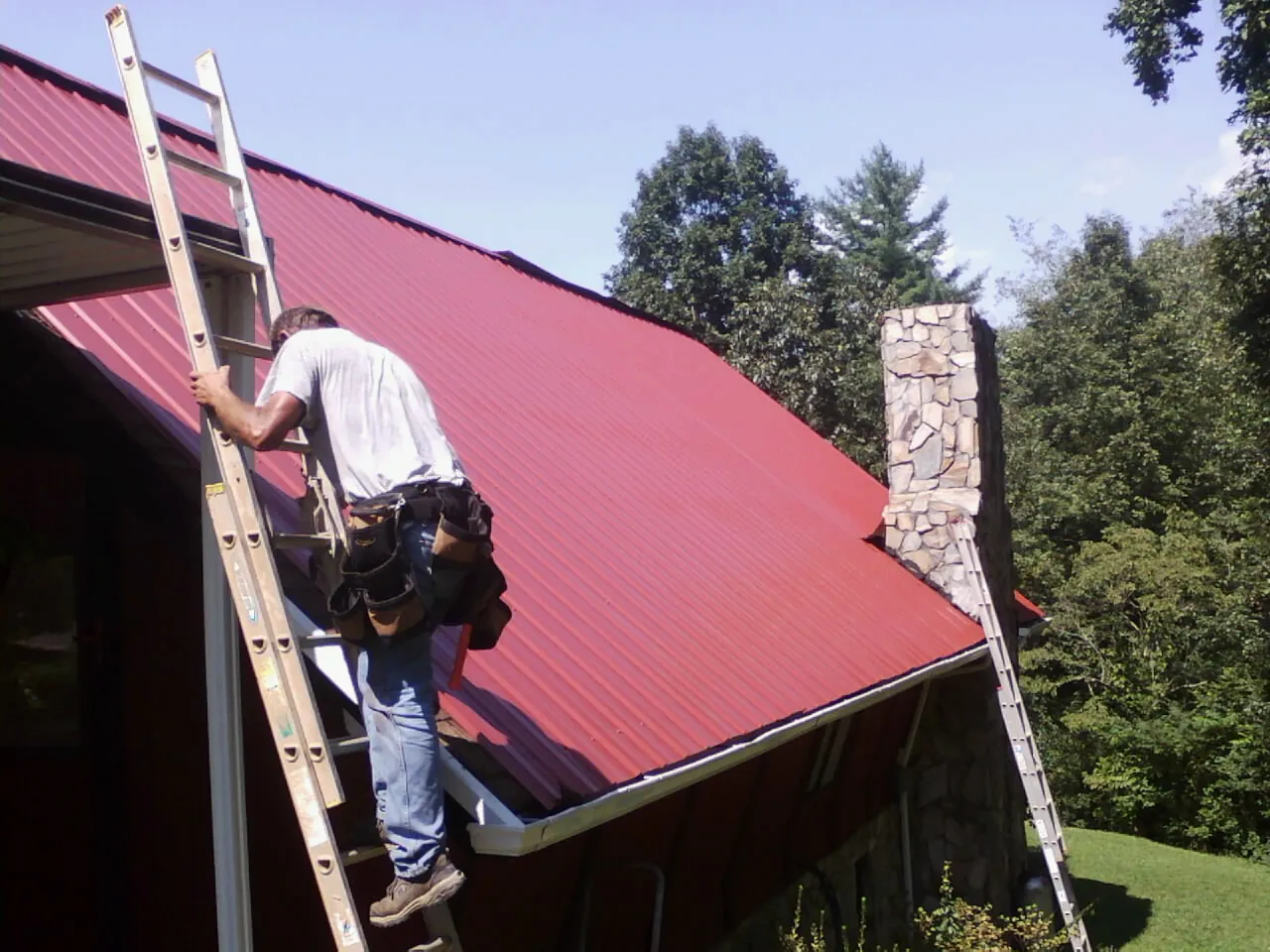 Roofer on a ladder beside a red metal roof and stone chimney — Soffit & Fascia Repair by Roofing Brothers in Jewett City